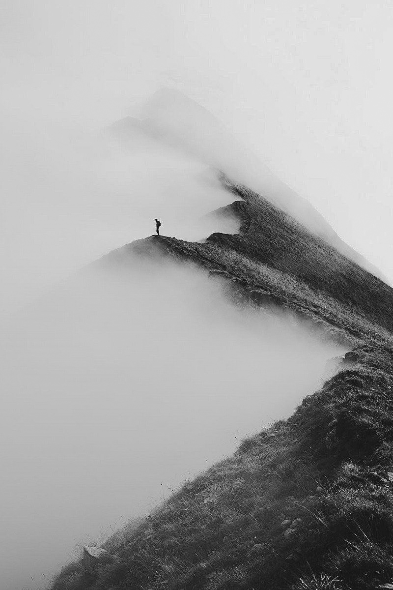 A lone figure standing on a narrow mountain ridge photographed in black and white, the slope falling away into cloud on either side.